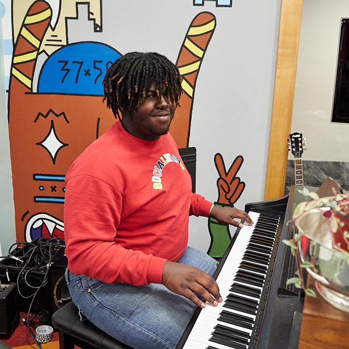 A teenager in a red sweatshirt playing the keyboard in a music room.